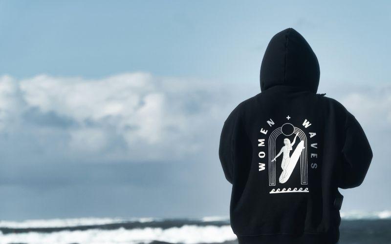 A person wearing a black hoodie with a large white women and waves logo placed on the back. A good example of how to choose the right logo placement on your custom hoodie.